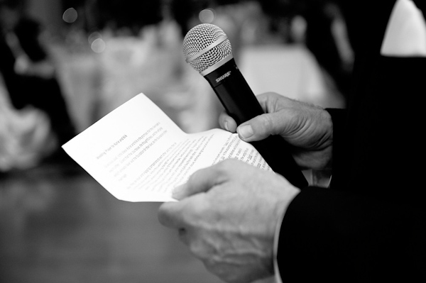 black and white photo - groomsman holding typed out speech and microphone while giving a toast at the reception -  photo by Houston based wedding photographer Adam Nyholt